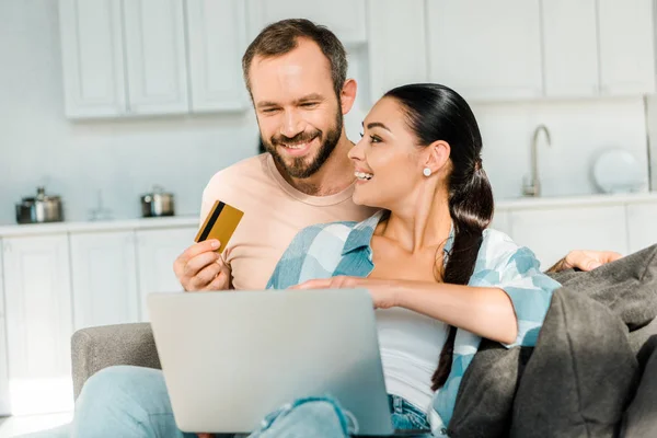 Красивий чоловік тримає кредитну картку, посміхаючись дружині, використовуючи ноутбук і здійснюючи онлайн-покупки вдома — стокове фото