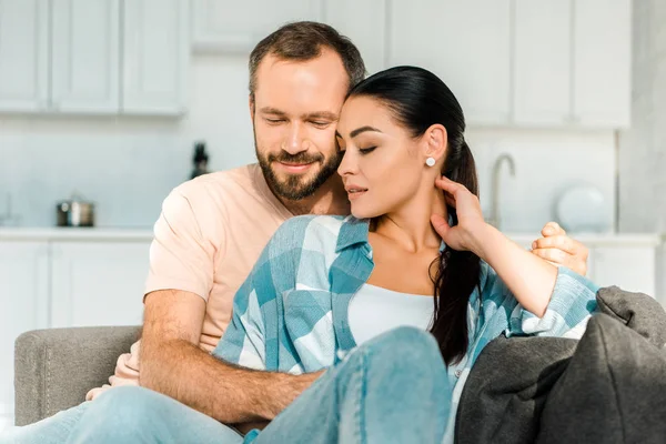 Handsome husband and beautiful wife sitting and embracing on couch at home — Stock Photo