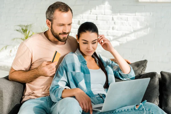 Marido guapo sosteniendo la tarjeta de crédito mientras que la esposa hermosa usando el ordenador portátil y haciendo compras en línea en el país - foto de stock