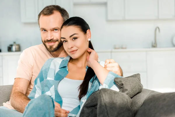 Feliz pareja mirando a la cámara, abrazando y sentado en el sofá en casa - foto de stock
