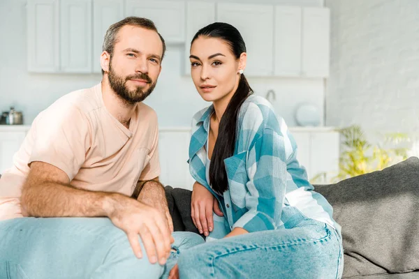 Beau couple aimant regardant la caméra et assis sur le canapé à la maison — Photo de stock