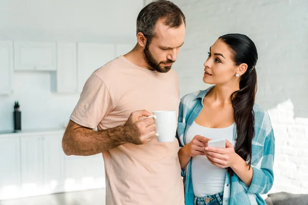 Beautiful wife using smartphone while handsome husband holding cup of coffee at home — Stock Photo
