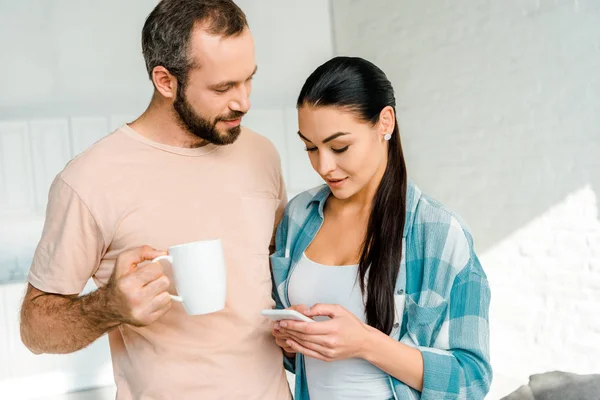 Beautiful brunette wife using smartphone while husband holding cup of coffee at home — Stock Photo