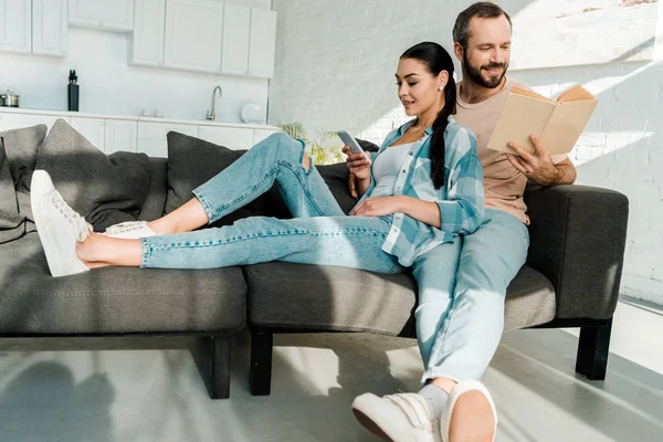 Lächelnde Ehefrau mit Smartphone, während Mann zu Hause auf Couch sitzt und Buch liest — Stockfoto