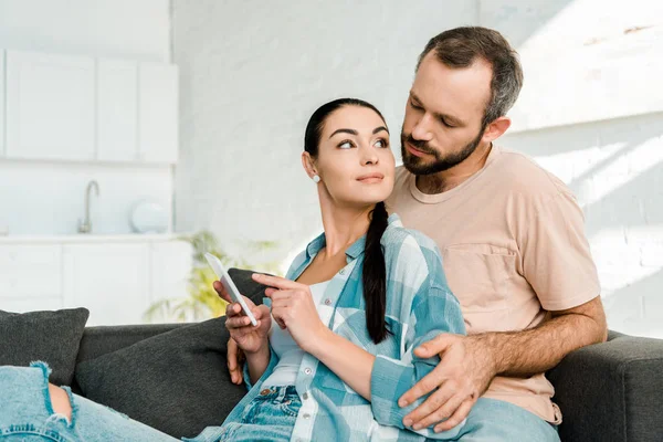 Casal atraente sentado no sofá e usando smartphone em casa — Fotografia de Stock