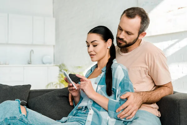 Husband and wife sitting on couch and using smartphone at home — Stock Photo