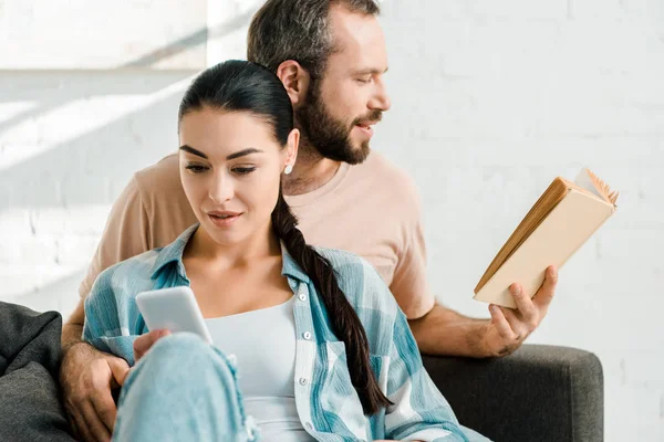 Atractiva esposa usando teléfono inteligente, mientras que el hombre sentado en el sofá y libro de lectura en casa - foto de stock