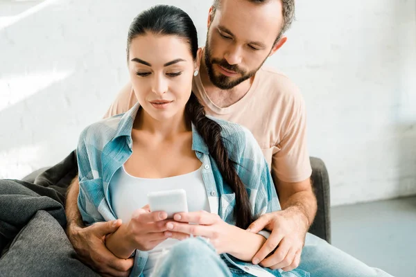 Attractive couple sitting on couch, hugging and using smartphone at home — Stock Photo