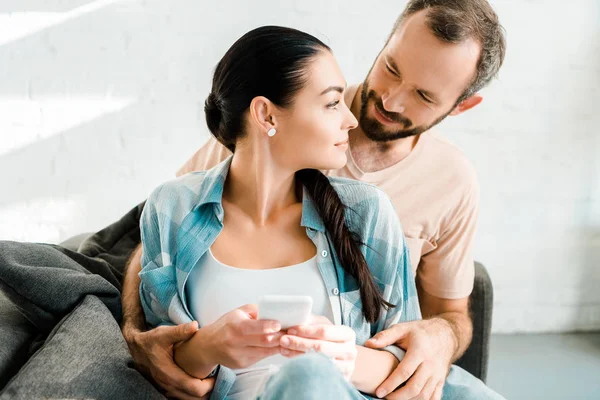 Loving couple sitting on couch, hugging and using smartphone at home — Stock Photo