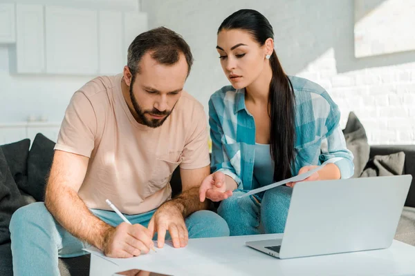 Fokussiertes Paar, das auf der Couch sitzt, Papiere füllt, Laptop benutzt und zu Hause plant — Stockfoto