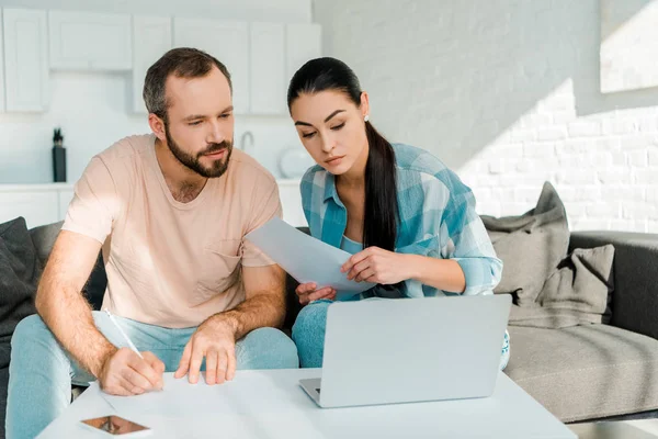 Concentrated couple sitting on couch, filling papers, using laptop and planning — Stock Photo