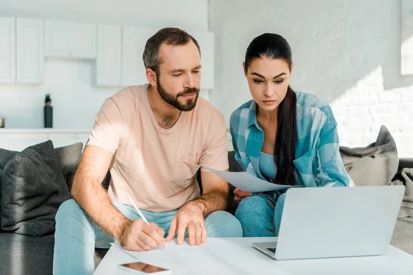 Serious couple sitting on couch, filling papers, using laptop and planning at home — Stock Photo