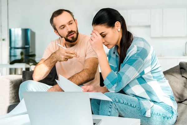 Pareja cansada sentada en el sofá, usando el ordenador portátil y teniendo problemas financieros en casa - foto de stock