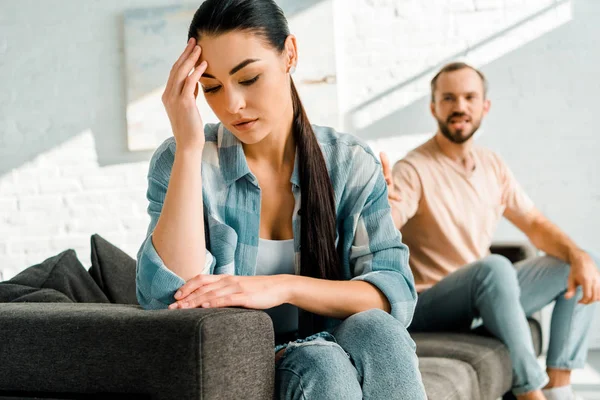 Femme stressée avec la main sur la tête assise au premier plan après s'être disputée avec son mari à la maison — Photo de stock