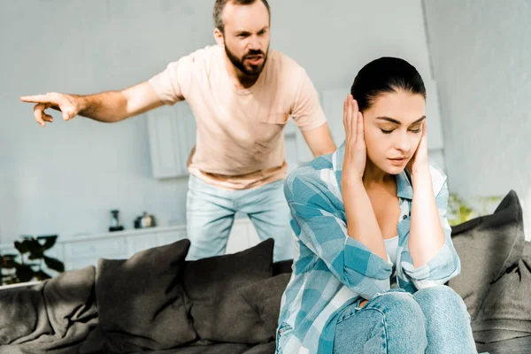 Enojado marido gritando a la esposa sentada en el sofá y cubriendo las orejas con las manos — Stock Photo