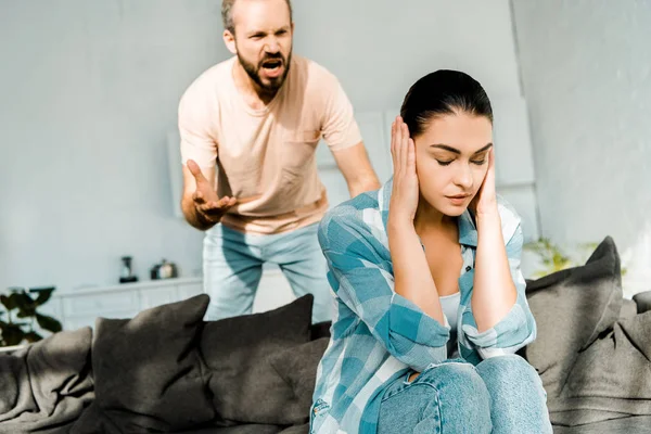 Enojado marido gritando a la esposa estresada sentado en el sofá y cubriendo las orejas con las manos - foto de stock