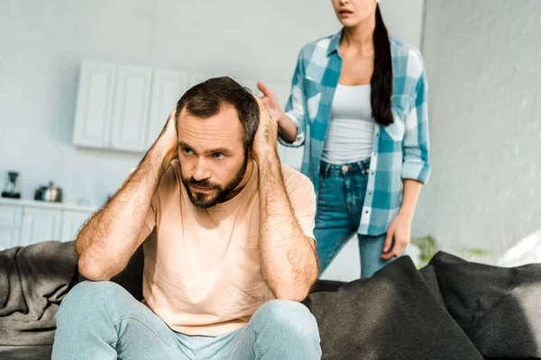 Estresado marido en primer plano cubriendo las orejas con las manos y tener una discusión con la esposa en casa - foto de stock