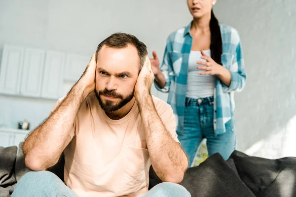 Marido zangado em primeiro plano cobrindo orelhas com as mãos e tendo discussão com a esposa em casa — Fotografia de Stock