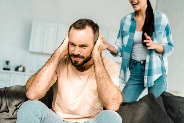Розчарований чоловік на передньому плані покриває вуха руками і має суперечки з дружиною вдома — стокове фото