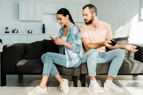 Dissatisfied husband looking at wife sitting on couch and using smartphone — Stock Photo