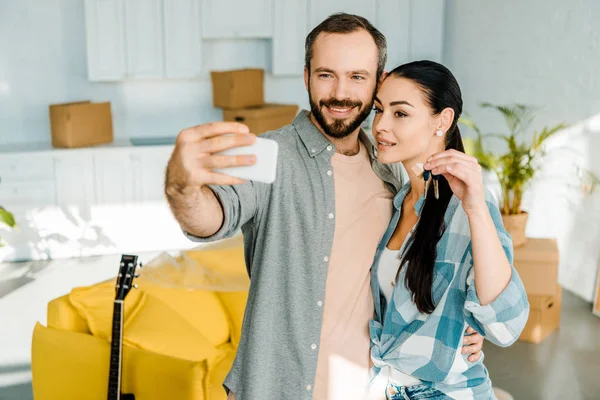 Mari souriant prenant selfie sur smartphone tandis que la femme tenant les clés de la nouvelle maison — Photo de stock