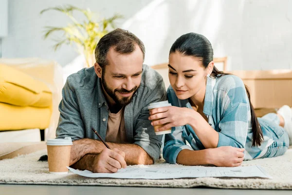 Mann und schöne Frau auf dem Boden liegend, Kaffee trinkend und zu Hause am Bauplan des neuen Hauses arbeitend, bewegendes Konzept — Stockfoto