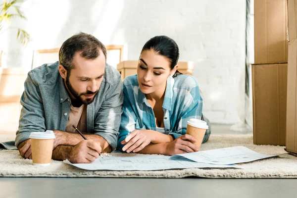 Handsome husband and wife lying on floor, drinking coffee and working on blueprints of new house at home, moving concept — Stock Photo