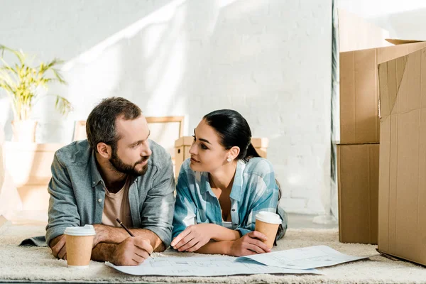 Mann und attraktive Frau auf dem Boden liegend, Kaffee trinkend und zu Hause an Entwürfen für neues Haus arbeitend, bewegendes Konzept — Stockfoto