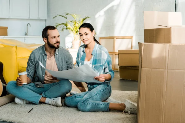 Sonrientes marido y mujer bebiendo café y sosteniendo el plano de la nueva casa, concepto en movimiento - foto de stock