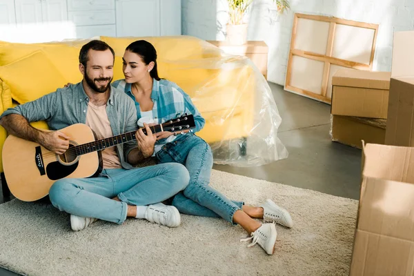 Красивый муж, сидящий на полу и играющий на акустической гитаре, пока жена слушает — стоковое фото
