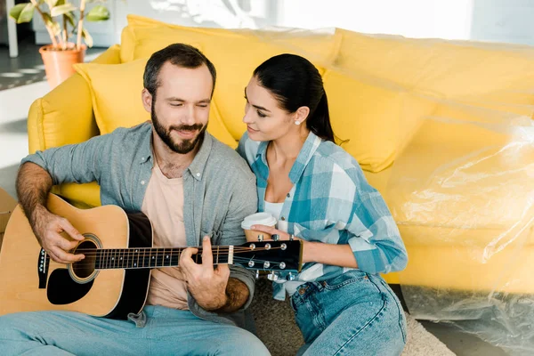 Чоловік сидить на підлозі і грає на акустичній гітарі, а красива дружина слухає — стокове фото