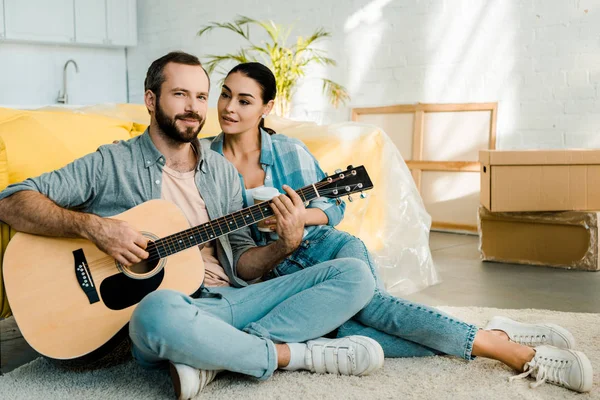 Marido bonito tocando guitarra acústica e olhando para a câmera enquanto esposa ouvindo — Fotografia de Stock