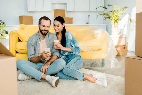 Улыбающаяся пара пьет кофе, используя смартфон и расслабляясь после упаковки для нового дома, движущаяся концепция — стоковое фото