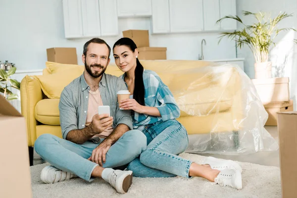 Couple boire du café, en utilisant un smartphone et se détendre après l'emballage pour la maison neuve, concept mobile — Photo de stock