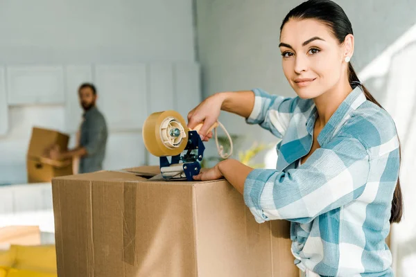 Wife packing cardboard box with scotch tape and husband behind, moving concept — Stock Photo