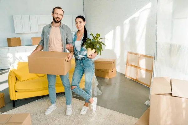 Husband carrying cardboard box and wife holding green plant while packing for into new house, moving concept — Stock Photo