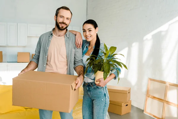 Marido que lleva la caja de cartón y la esposa que sostiene la planta verde mientras que embala para la casa nueva, concepto móvil - foto de stock