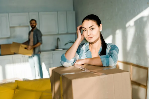 Wife leaning on cardboard box with husband packing for new house on background, moving concept — Stock Photo