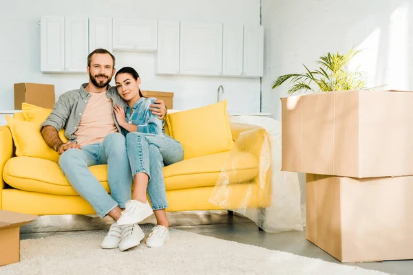 Smiling husband and wife embracing and sitting on couch while packing for new house, moving concept — Stock Photo