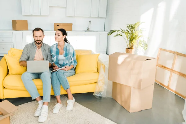 Pareja concentrada sentada en el sofá, utilizando el ordenador portátil y la planificación de la reubicación a la nueva casa, el concepto de movimiento - foto de stock