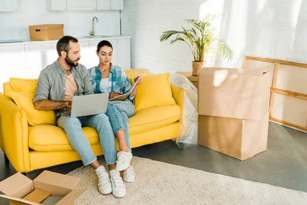 Beau couple assis sur le canapé, en utilisant un ordinateur portable et la planification de déménagement dans une nouvelle maison, concept de déménagement — Photo de stock