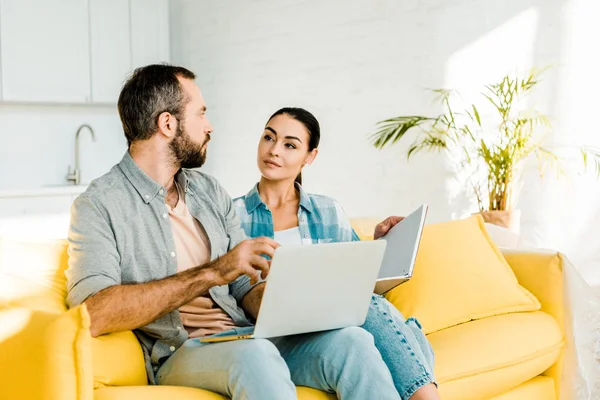 Bonito marido usando laptop enquanto bela esposa segurando notebook e sentado no sofá — Fotografia de Stock