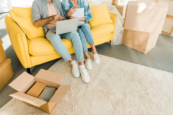 Обрезанный вид на пару, сидящую на диване, с помощью ноутбука и планирования переселения в новый дом, движущаяся концепция — стоковое фото