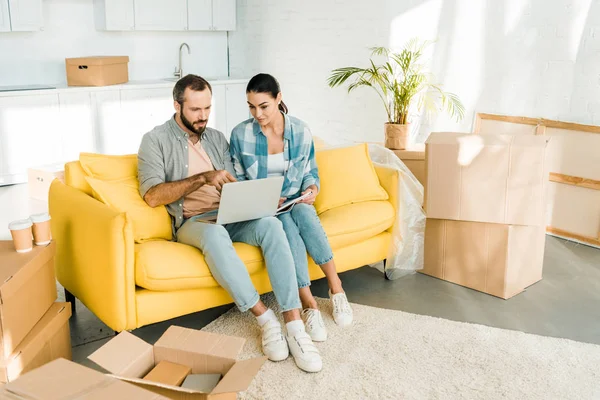 Целенаправленная пара, сидящая на диване, используя ноутбук и планируя переселение в новый дом, движущаяся концепция — стоковое фото