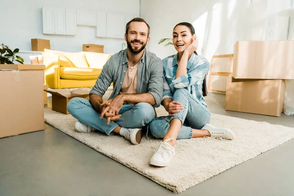 Marido y mujer sentados en el suelo y sonriendo después de empacar para una nueva casa, concepto en movimiento - foto de stock