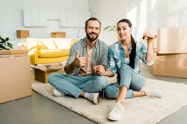 Marido haciendo pulgares hacia arriba signo mientras que la esposa sosteniendo las llaves de la casa nueva, concepto en movimiento - foto de stock