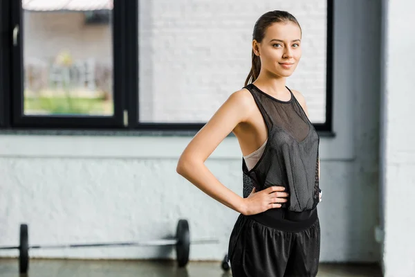 Attraente sportiva vestibilità in nero con le mani sui fianchi guardando la fotocamera in palestra — Foto stock