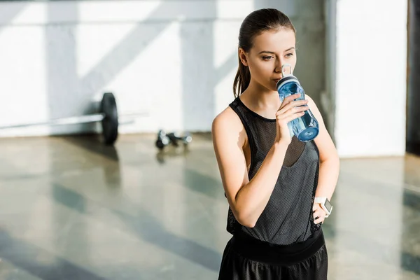 Красивая концентрированная спортсменка пьет воду из спортивной бутылки в спортзале — стоковое фото