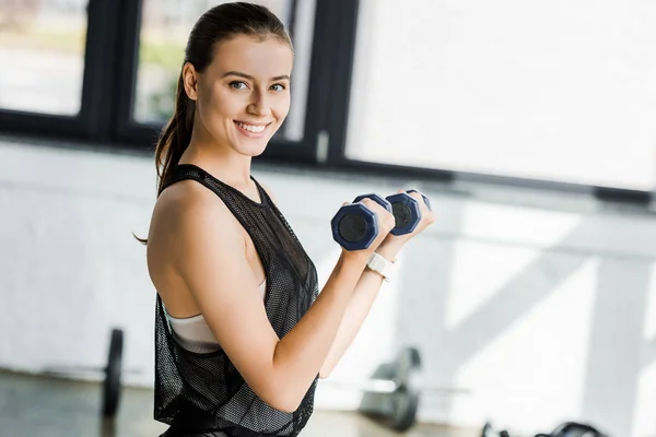 Beautiful smiling sportswoman looking at camera and training with dumbbells at sports center — Stock Photo