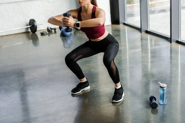 Обрезанный вид спортивной спортсменки, делающей упражнения на корточках с гантелями в фитнес-центре — стоковое фото